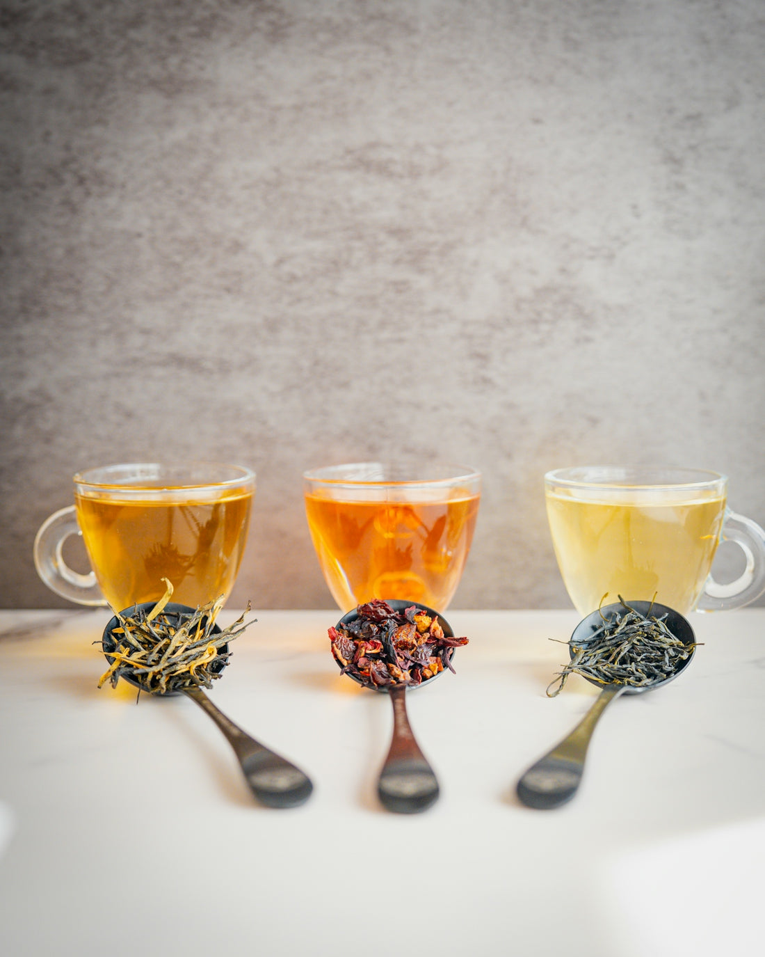 Three glass cups of tea with matching spoons of loose tea leaves