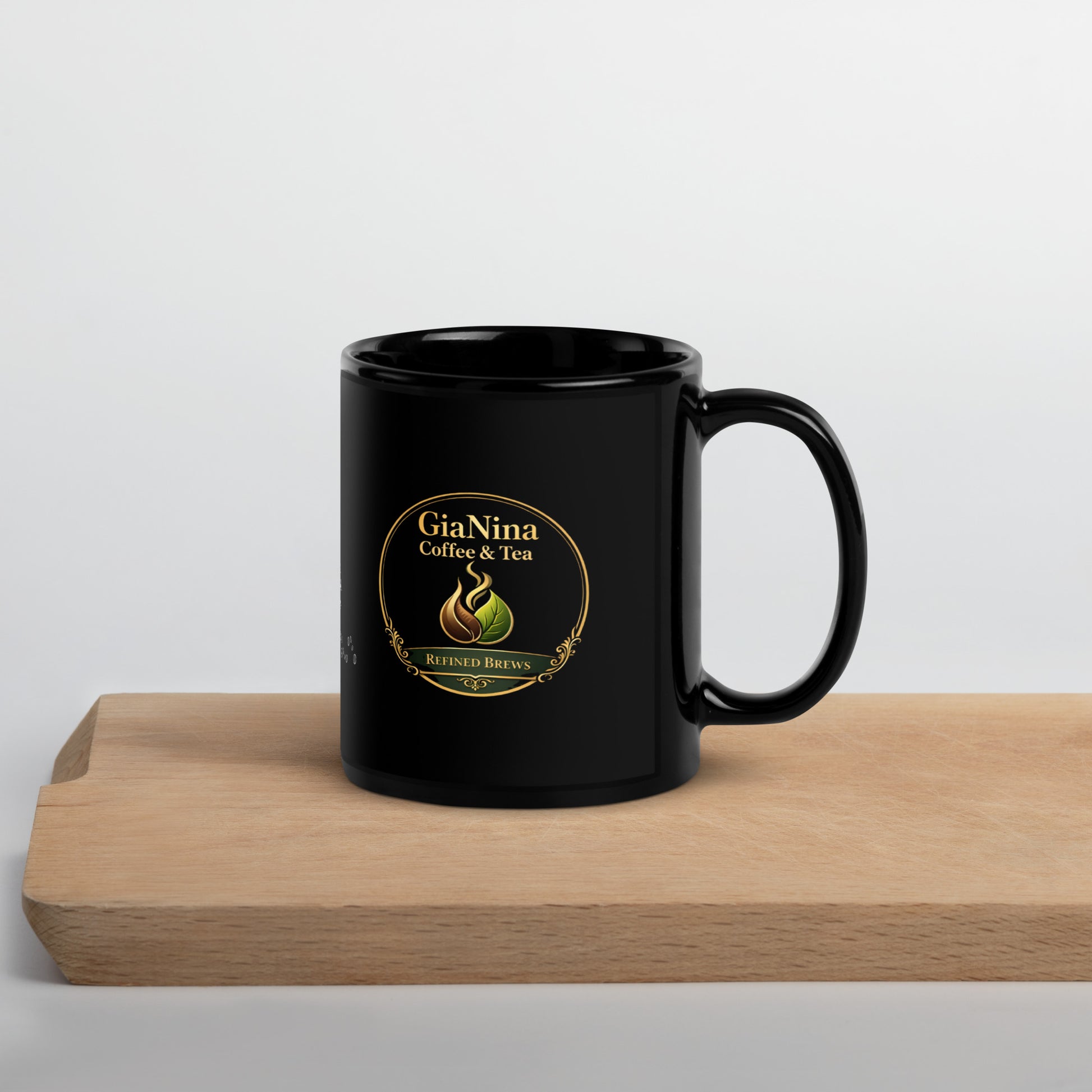 A black ceramic coffee mug with a gold and green GiaNina Coffee & Tea logo, displayed on a light wooden board against a neutral light gray background.