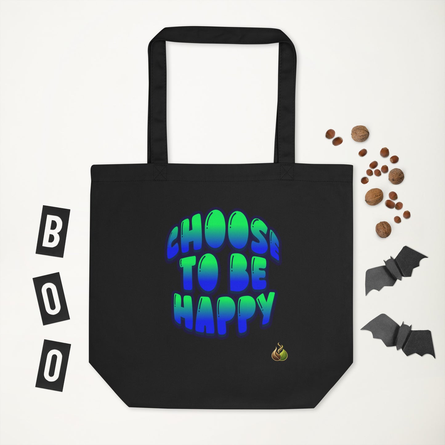 A black canvas tote bag with the phrase 'CHOOS TO BE HAPPY' printed in a blue-to-green gradient bubble font. The bag is displayed on a white background with Halloween-themed flat-lay props, including 'BOO' letter tiles, mini chocolate chips, and paper bats.