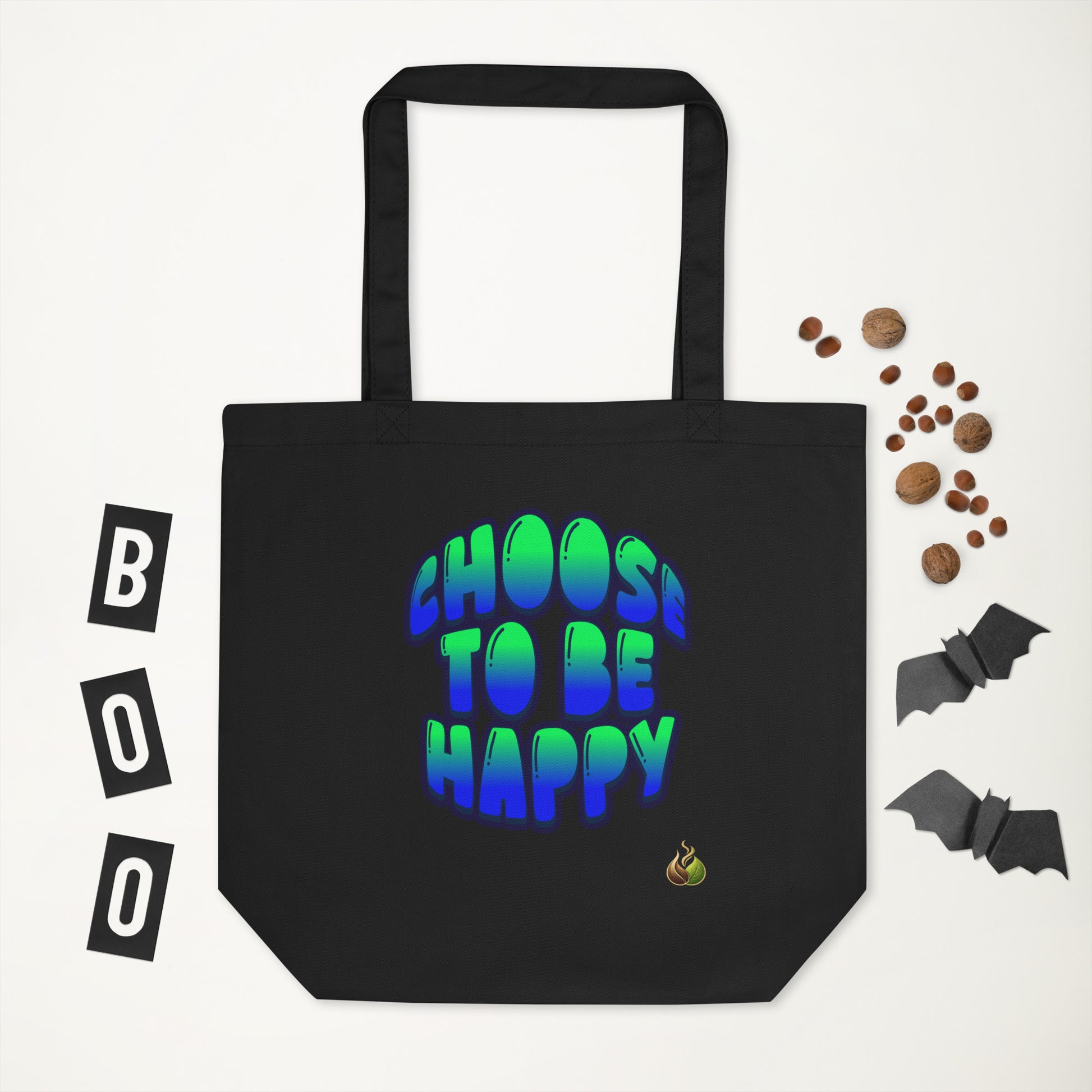 A black canvas tote bag with the phrase 'CHOOS TO BE HAPPY' printed in a blue-to-green gradient bubble font. The bag is displayed on a white background with Halloween-themed flat-lay props, including 'BOO' letter tiles, mini chocolate chips, and paper bats.