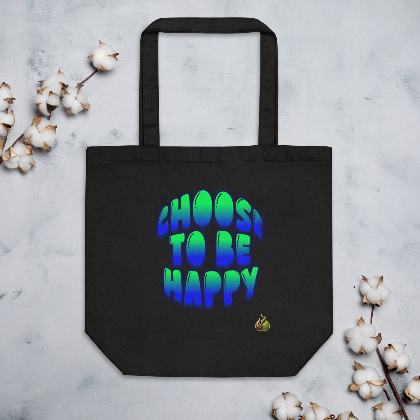 A black canvas tote bag with the phrase 'CHOOS TO BE HAPPY' printed in a vibrant green-to-blue gradient bubble font. The bag is displayed flat on a light gray marble surface, styled with decorative cotton bolls in the corners.