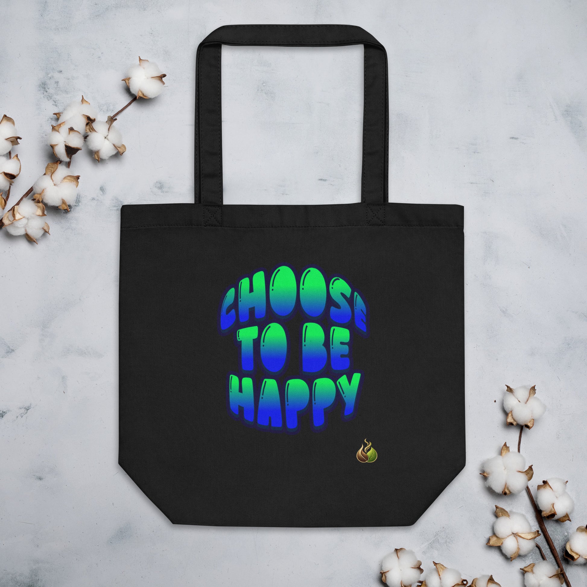 A black canvas tote bag with the phrase 'CHOOS TO BE HAPPY' printed in a vibrant green-to-blue gradient bubble font. The bag is displayed flat on a light gray marble surface, styled with decorative cotton bolls in the corners.