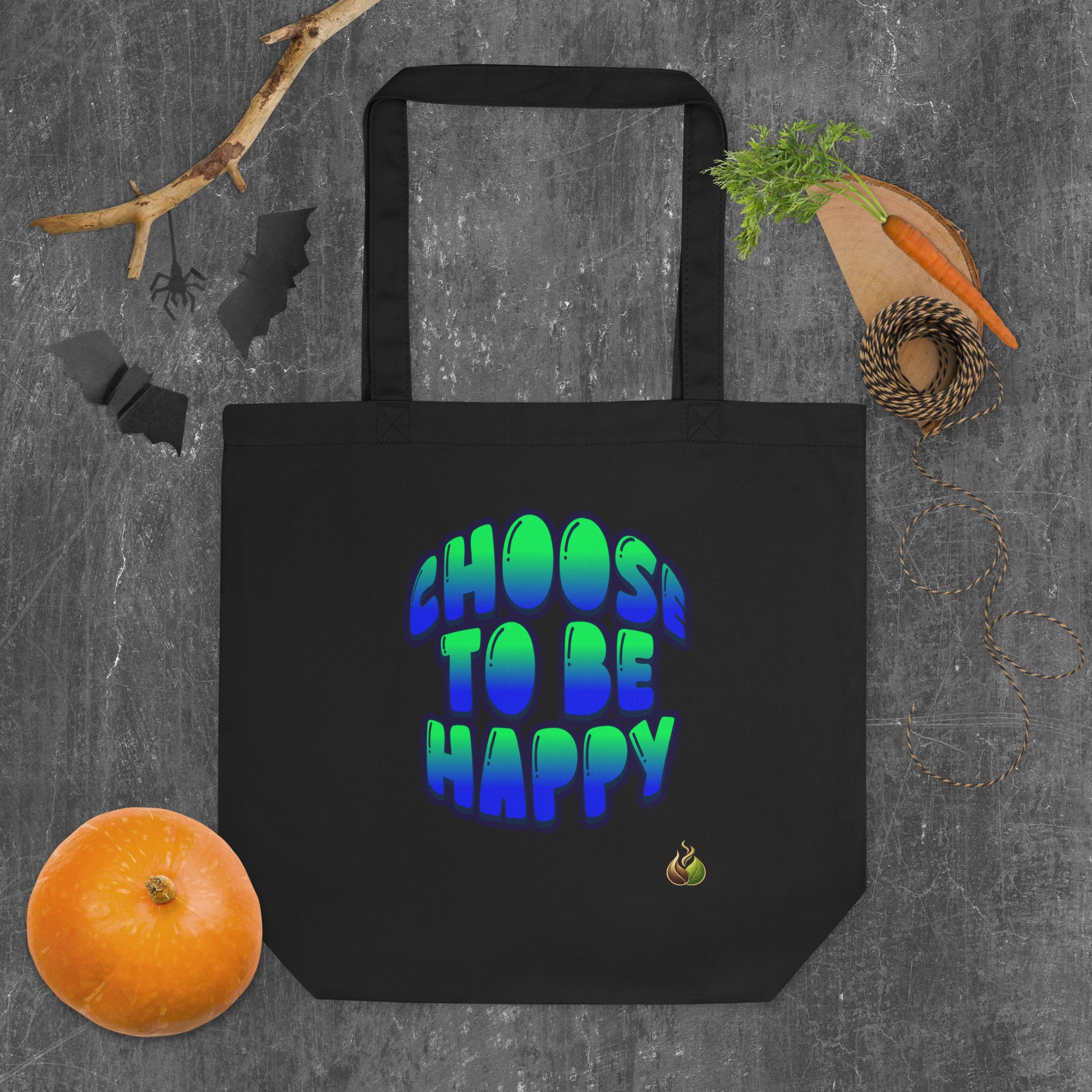 A black canvas tote bag with the phrase 'CHOOS TO BE HAPPY' printed in a blue-to-green gradient bubble font. The bag is displayed in a flat-lay arrangement on a textured gray background, surrounded by Halloween-themed props including a small orange pumpkin, a wooden carving board with a carrot, black bat cutouts, and a spider decoration.