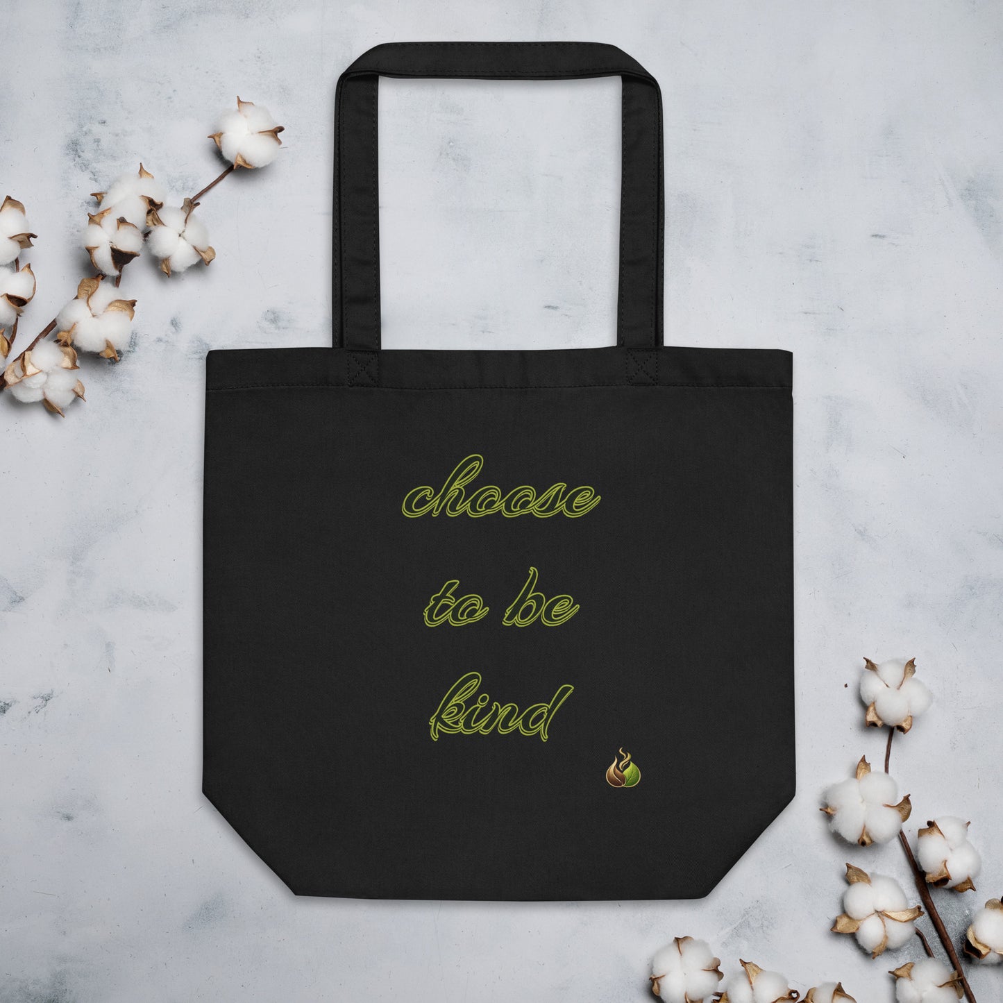 A black canvas tote bag with the phrase 'choose to be kind' embroidered in light green script, displayed flat on a light gray marble surface with decorative cotton bolls around it.