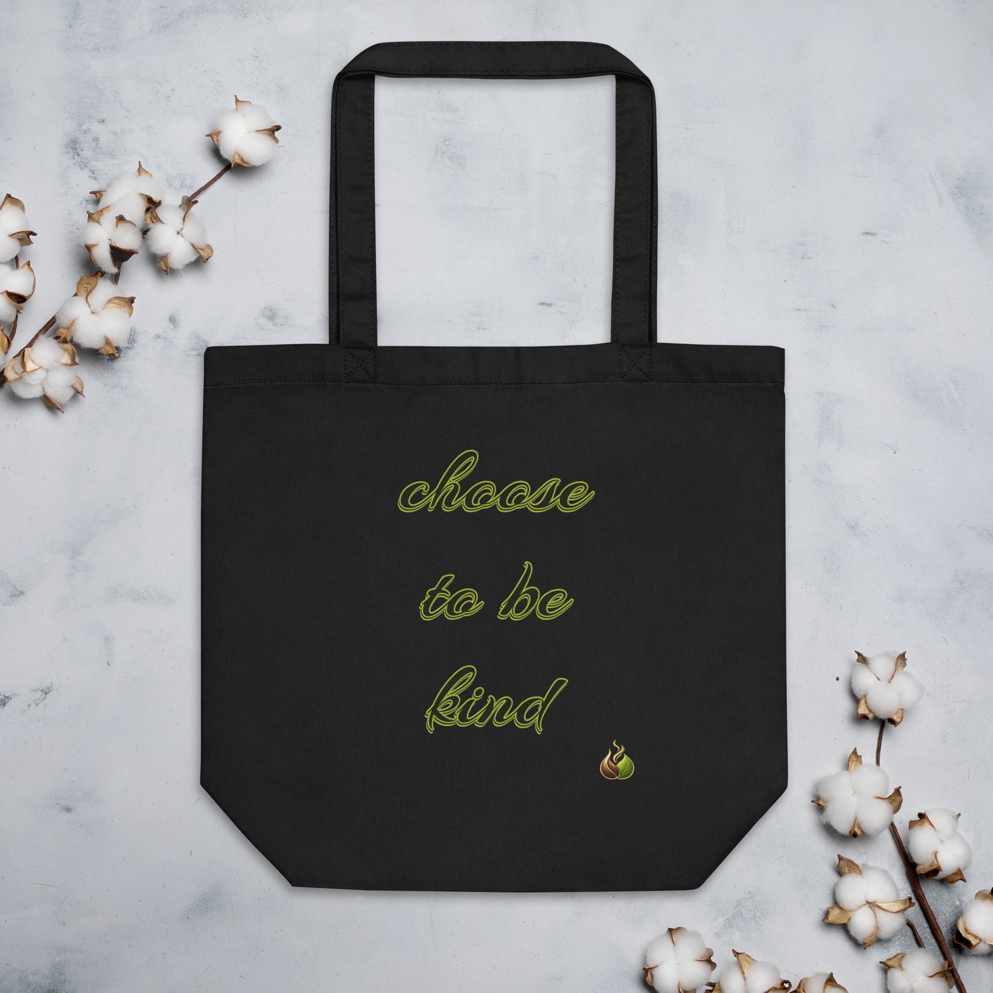 A black canvas tote bag with the phrase 'choose to be kind' embroidered in light green script, displayed flat on a light gray marble surface with decorative cotton bolls around it.