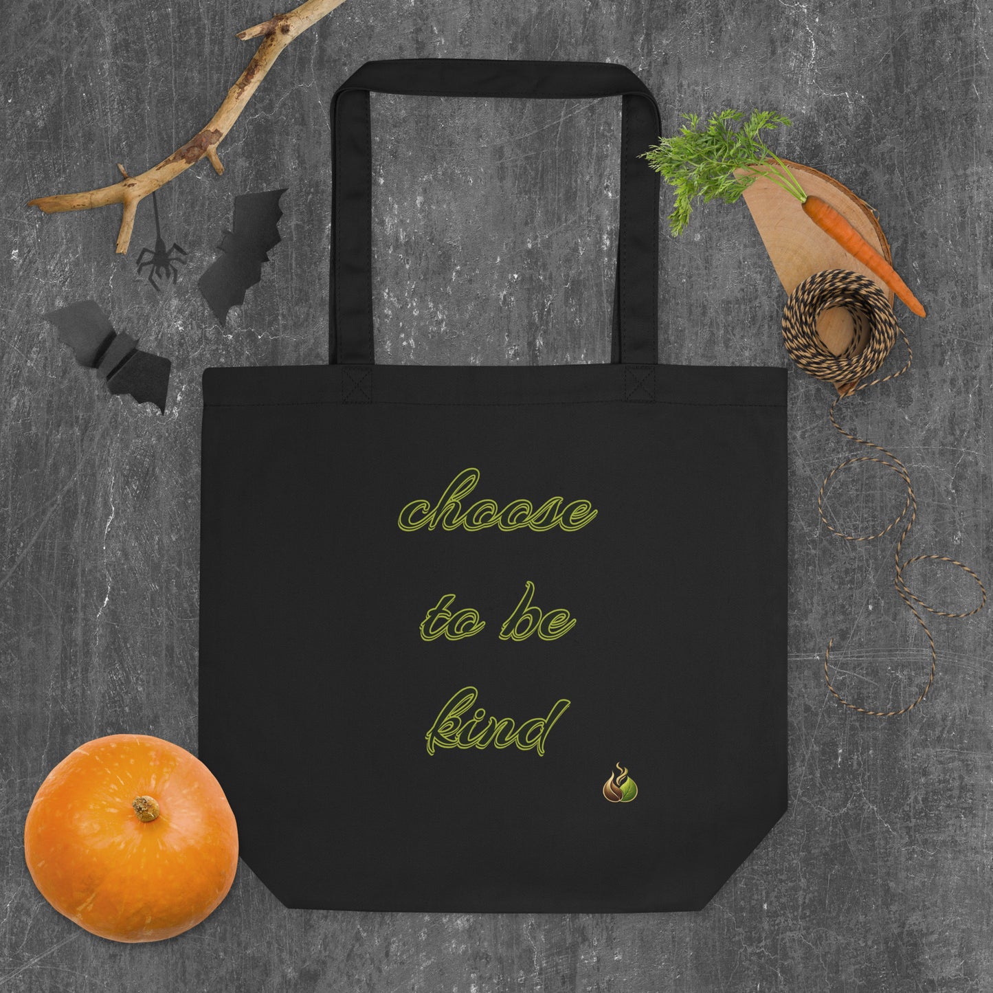 A black canvas tote bag with the phrase 'choose to be kind' printed in a yellow-green script, styled in a flat-lay arrangement on a dark gray textured background with autumn-themed props including a small orange pumpkin, a carrot, a pine sprig, and black bat silhouettes.