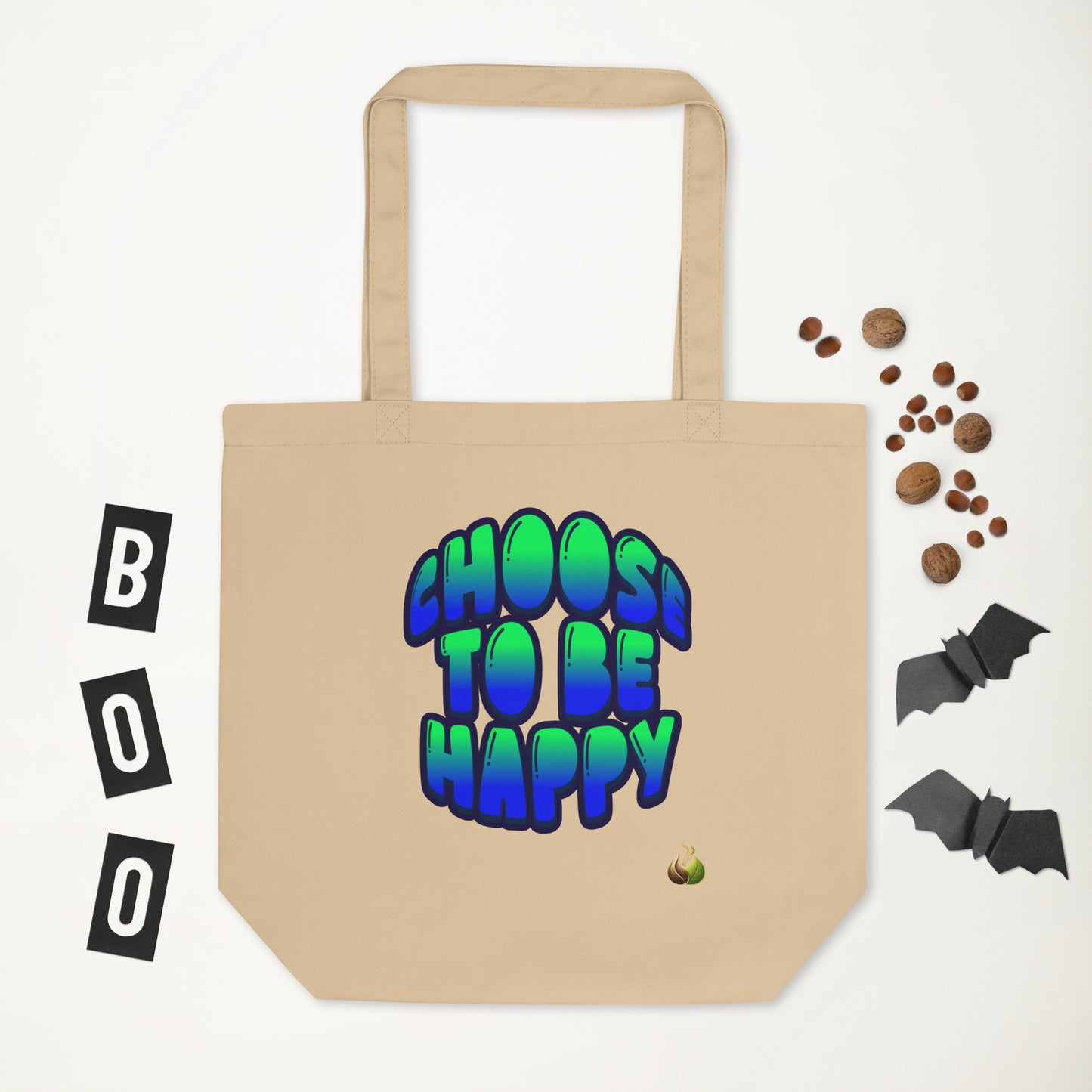 A beige canvas tote bag with the phrase 'CHOOS TO BE HAPPY' printed in a blue-to-green gradient bubble font with black outlines. The bag is displayed on a white background with Halloween-themed flat-lay props, including 'BOO' letter tiles, candy corn, and black bat cutouts.
