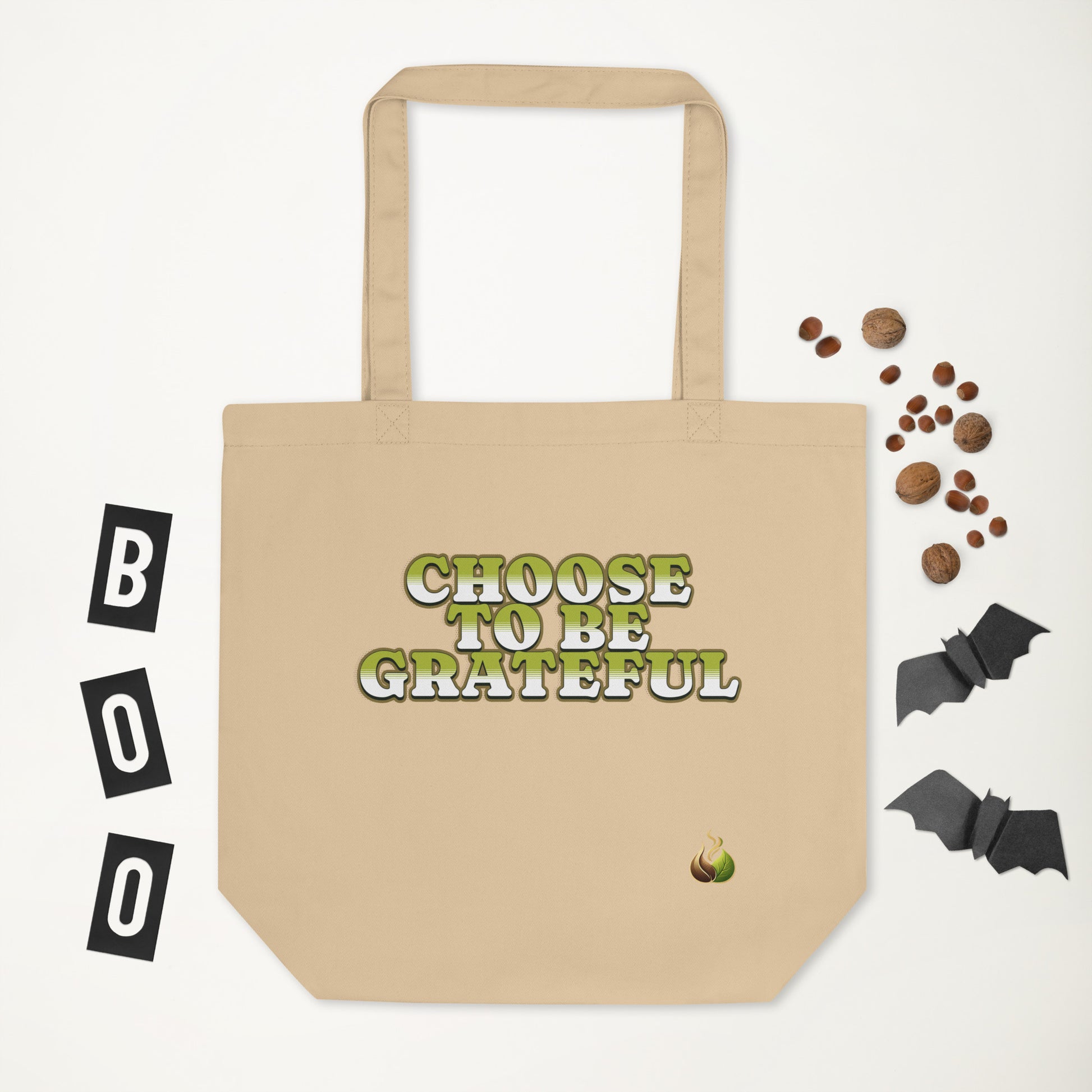 A beige canvas tote bag with the phrase 'CHOOSE TO BE GRATEFUL' printed in a green and white gothic font on the front. The bag is displayed on a clean white background with Halloween-themed flat-lay props, including three black blocks spelling 'BOO' on the left, scattered brown candies on the upper right, and two black bat cutouts on the lower right.