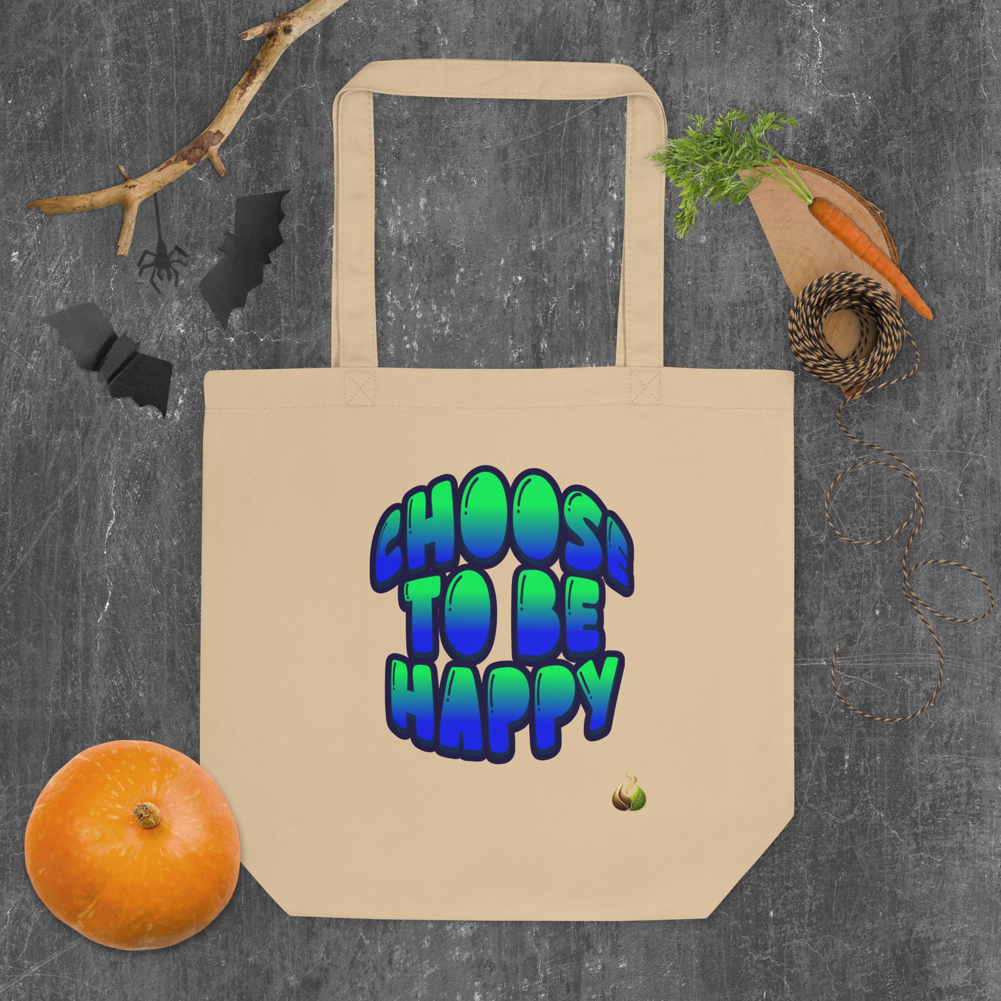 A beige canvas tote bag with the phrase 'CHOOS TO BE HAPPY' printed in a colorful 3D bubble font. The bag is displayed in a flat-lay arrangement on a dark gray textured background, surrounded by Halloween-themed props including a small orange pumpkin, a roll of twine, a carrot on a wooden board, and black bat silhouettes.