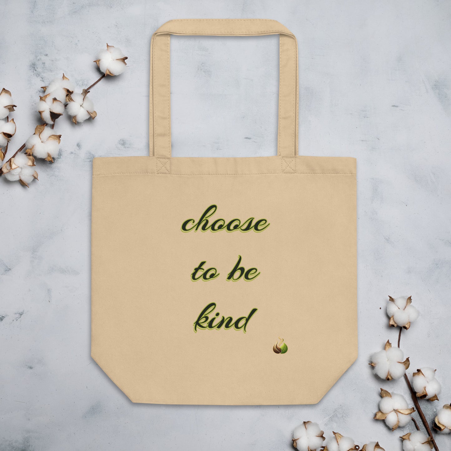 A beige canvas tote bag with the phrase 'choose to be kind' printed in green cursive text, styled on a light gray marble surface with cotton bolls around it.