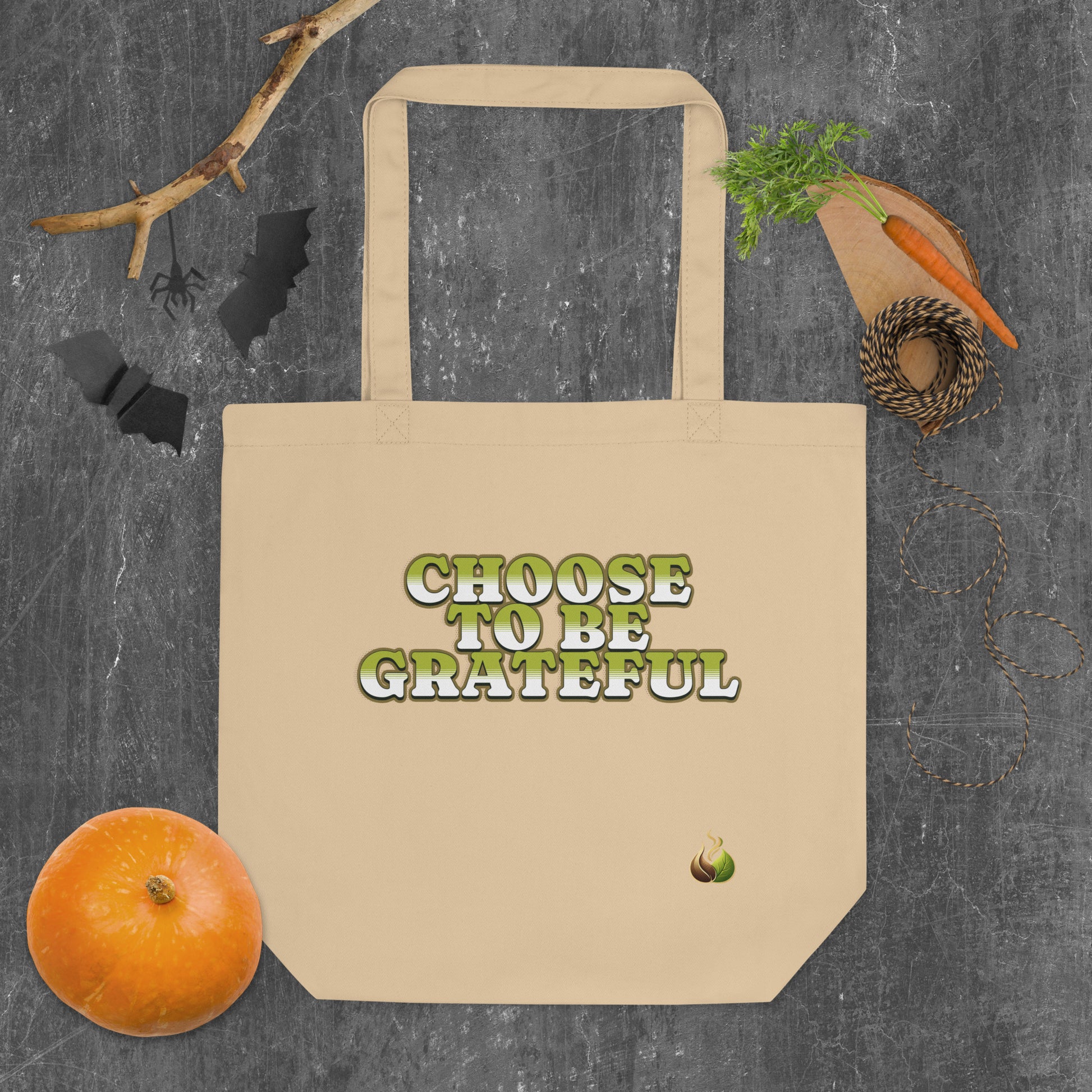 A beige canvas tote bag with the phrase 'CHOOSE TO BE GRATEFUL' printed on the front in green letters with a white outline. The bag is displayed in a flat-lay arrangement on a dark gray textured background, surrounded by seasonal props including a small orange pumpkin, a wooden cutting board with a carrot, a spool of twine, and black bat silhouettes.