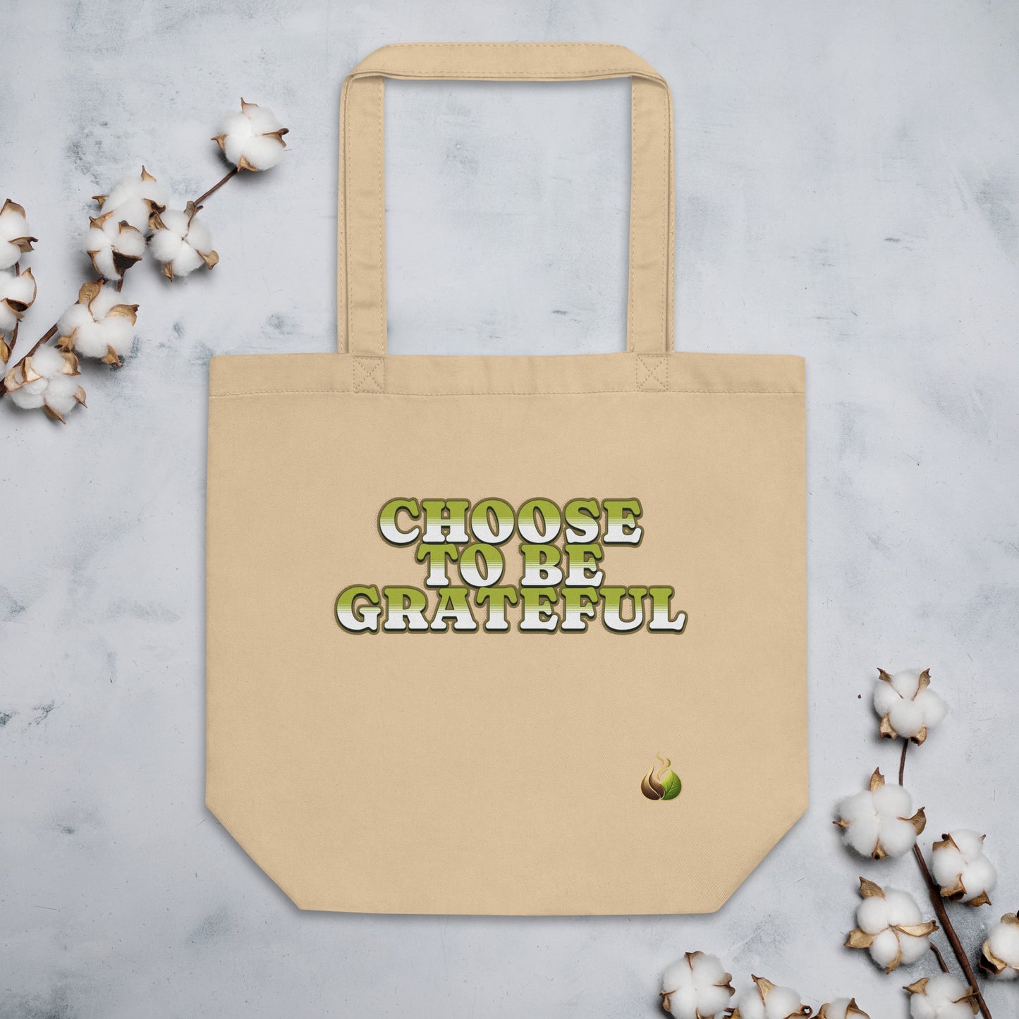 A beige canvas tote bag with the phrase 'CHOOSE TO BE GRATEFUL' printed on the front in green and white bubble letters, styled on a light gray marble surface with cotton bolls around it.