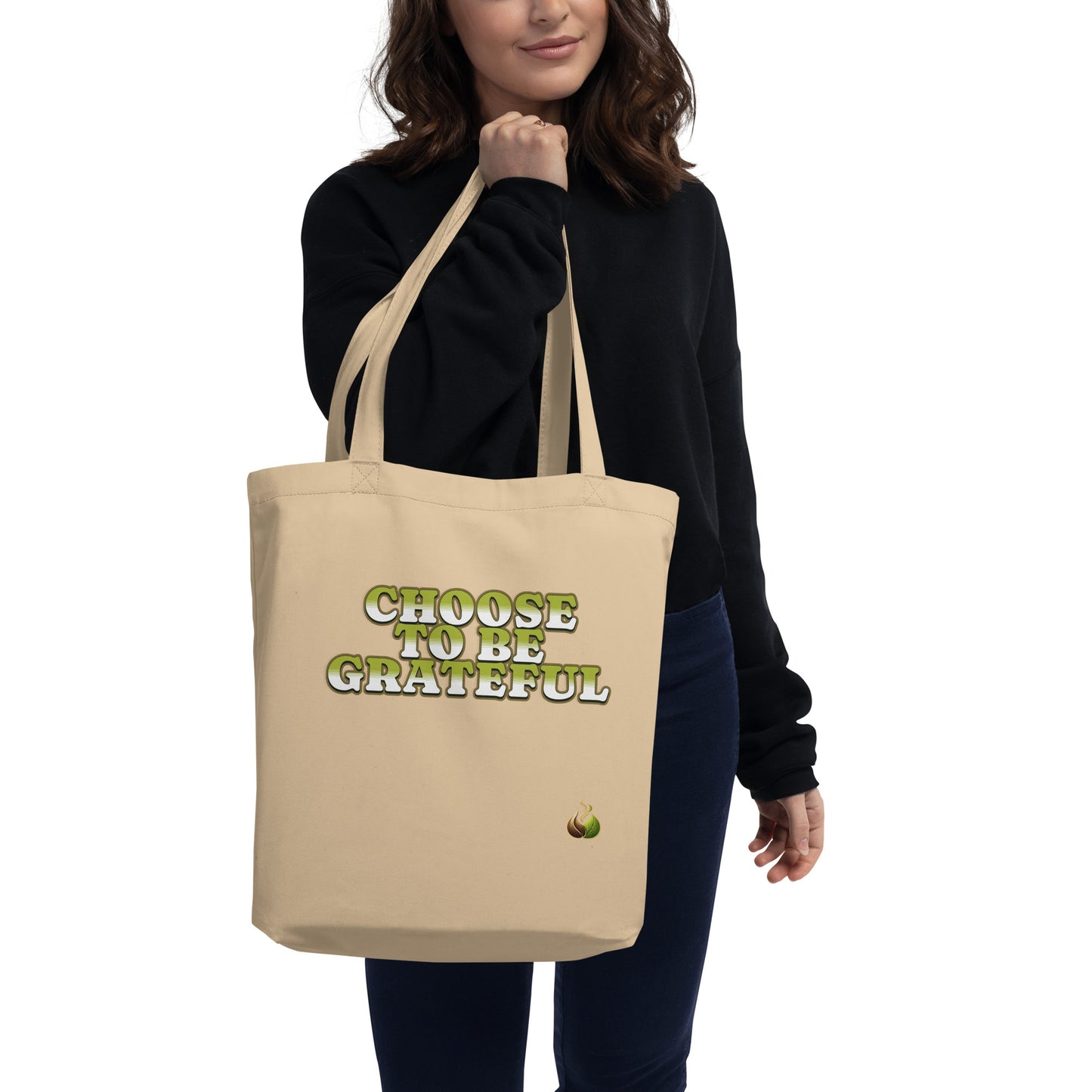 A woman wearing a black sweatshirt and dark blue jeans holds a beige canvas tote bag with the text 'CHOOSE TO BE GRATEFUL' in green and gold lettering, against a plain white background.